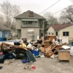 Piles of discarded furniture, cardboard, and household items outside a cluttered house.