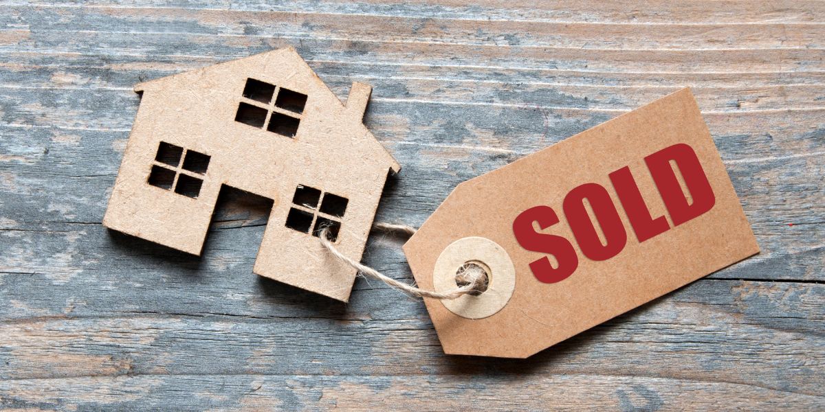 Cardboard house shape with a 'Sold' tag attached, placed on a wooden surface.