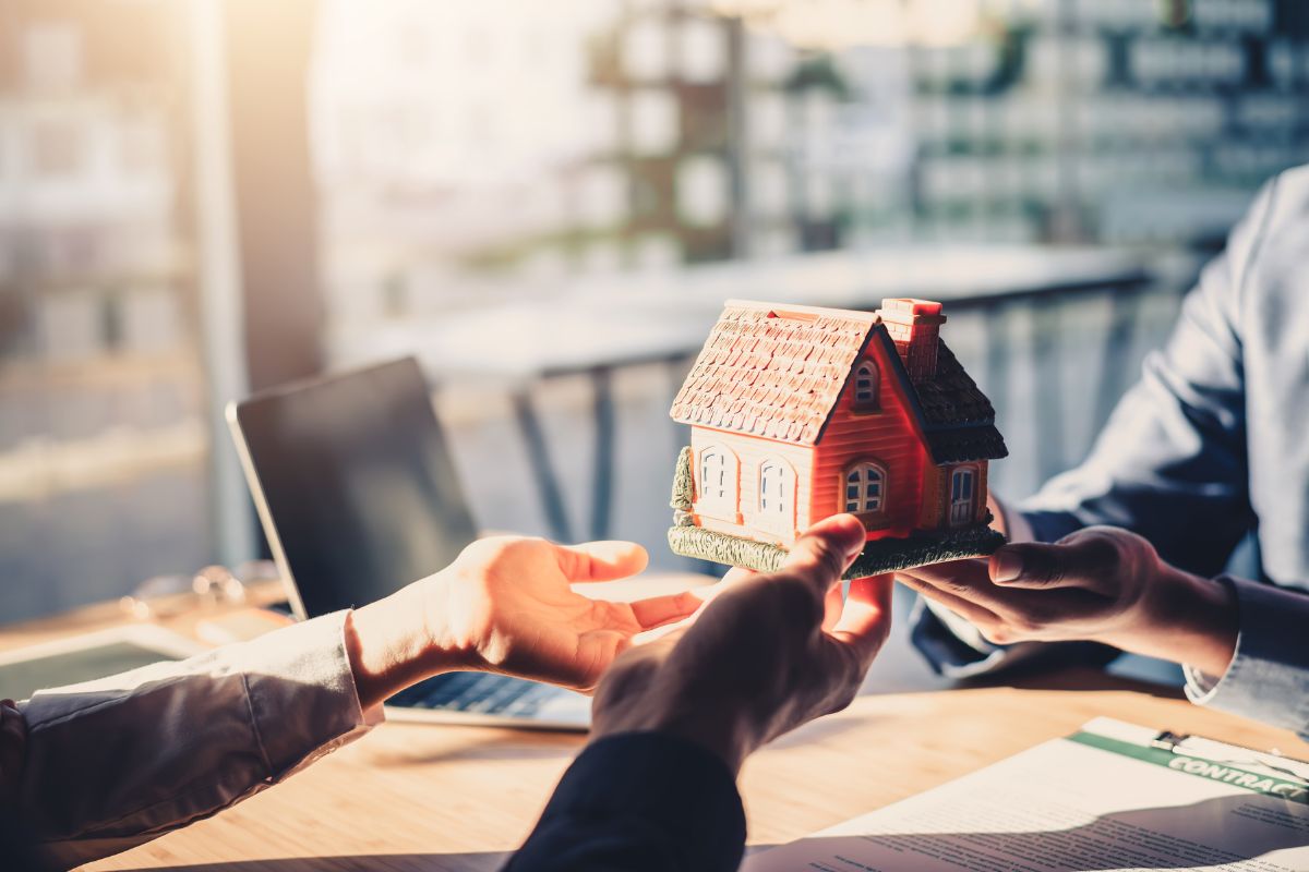 Hands exchanging a model house over a table with documents, symbolizing a seamless home sale process facilitated by REvitalize Property Solutions for a quick and hassle-free transaction.