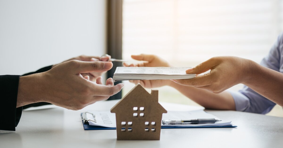 Two people exchanging paperwork during a real estate transaction with a small wooden house model in the foreground. One person is handing documents to another across a desk, symbolizing the hassle-free process of selling a home without the traditional complications.
