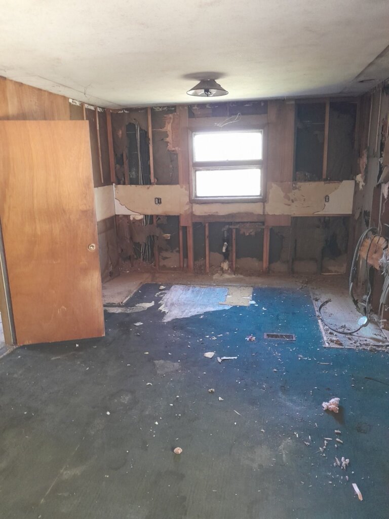 A room stripped of damaged cabinets with pipes and electrical visible from missing walls.