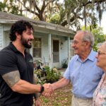 Jesse from Synergy Buys Houses shaking hands with a seller couple outside a distressed inherited Florida home with weathered siding, an aging roof, and an overgrown yard.