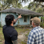 A weathered single-story Jacksonville home with an overgrown yard, a homeowner talking with a buyer on the front walk