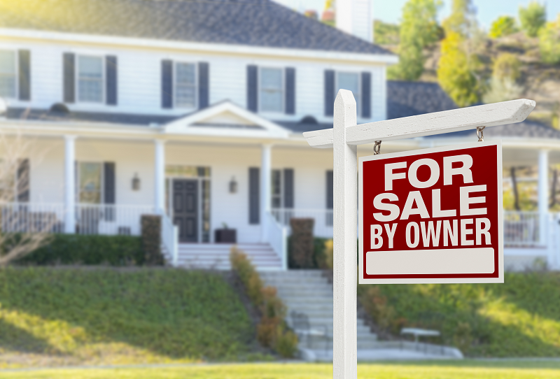 A red and white "For sale by Owner" sign with a house for sale in the background