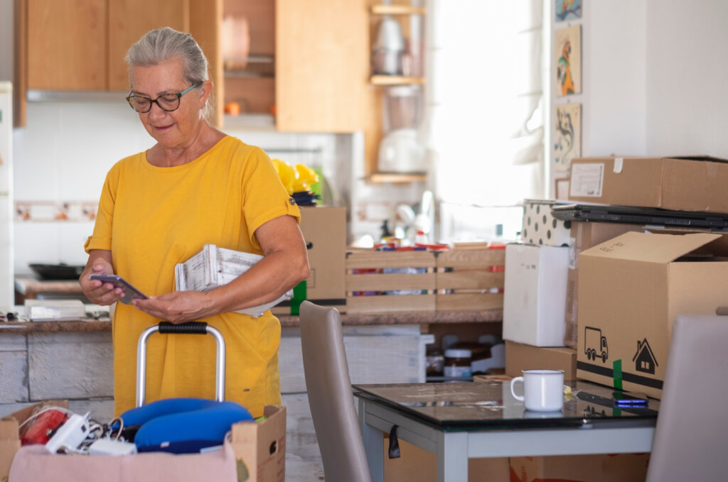 Woman looks at smart phone while holding a small white box. She's surrounded by moving supplies.
