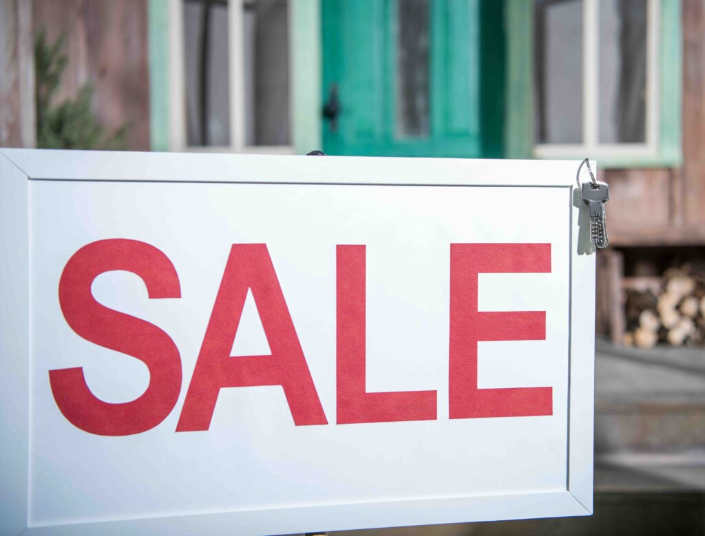 Photo of a closeup of a for sale sign in front of a rundown house.