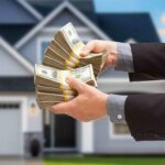 A photo of hands holding cash in front of a house to illustrate an article about realtor vs cash sale transaction.