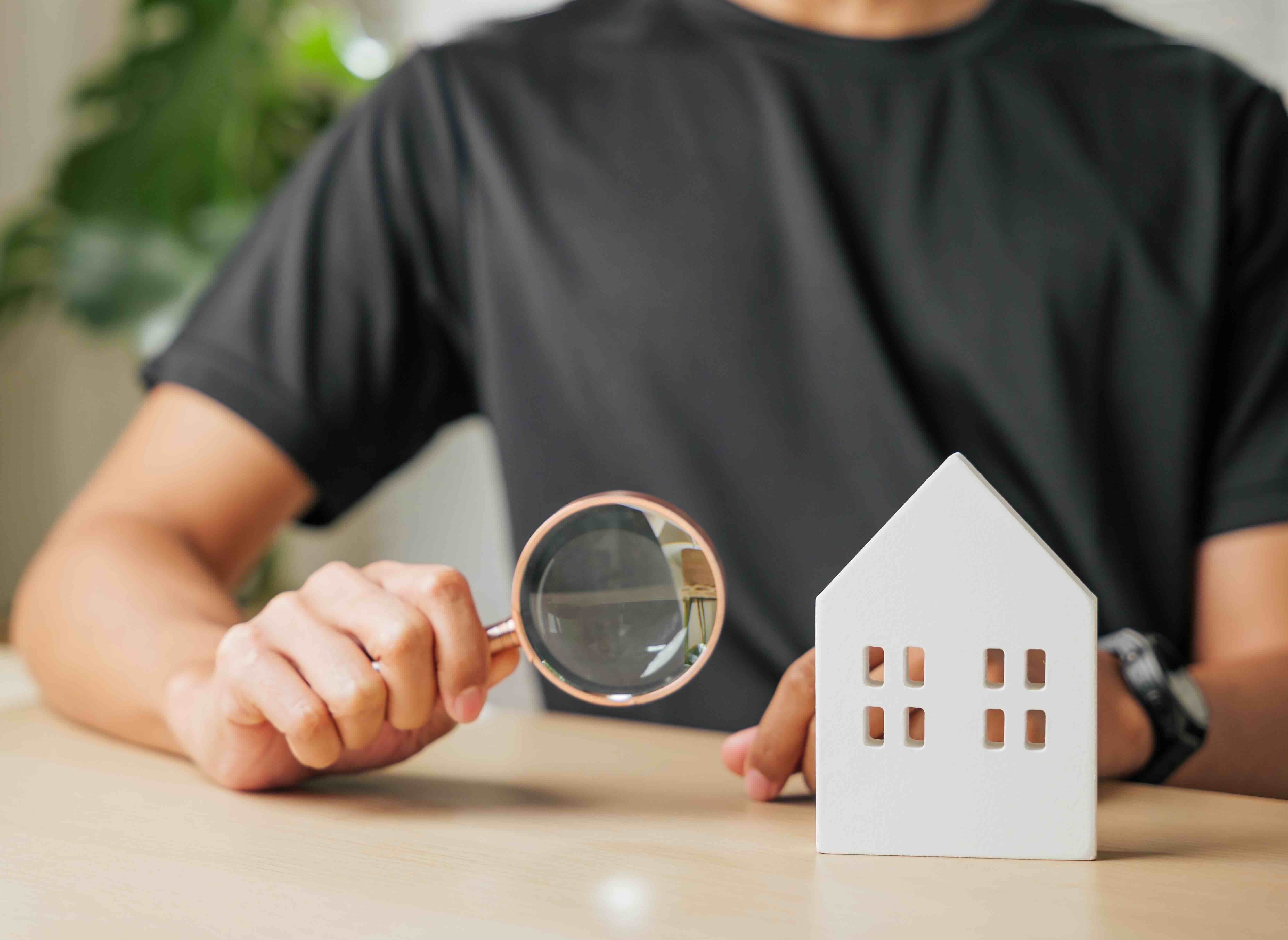 A photo of a man holding a magnifying glass to a small house, representing a home inspection.