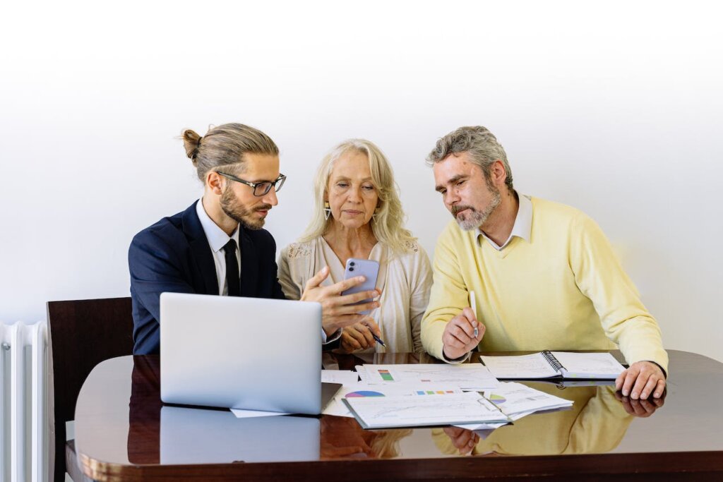 An older couple sitting and looking at the phone of a younger man while papers and a laptop are on the desk.