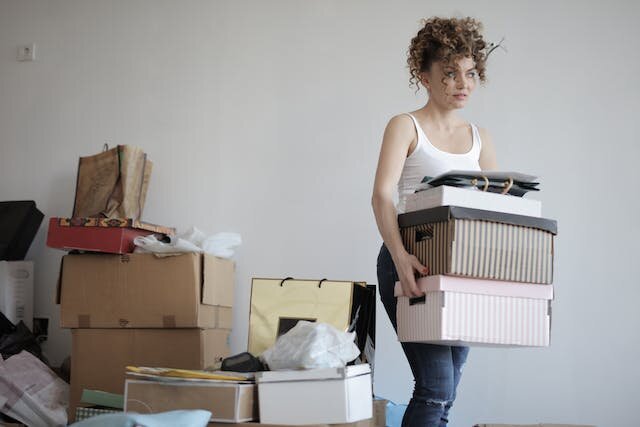 Decluttering is a great way to start prepping your home, followed by proper staging. Alt text: Woman holding stacked boxes next to a pile of clutter
