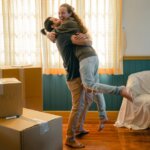 Two people embrace joyfully in a room with moving boxes, indicating they are moving out before selling their home