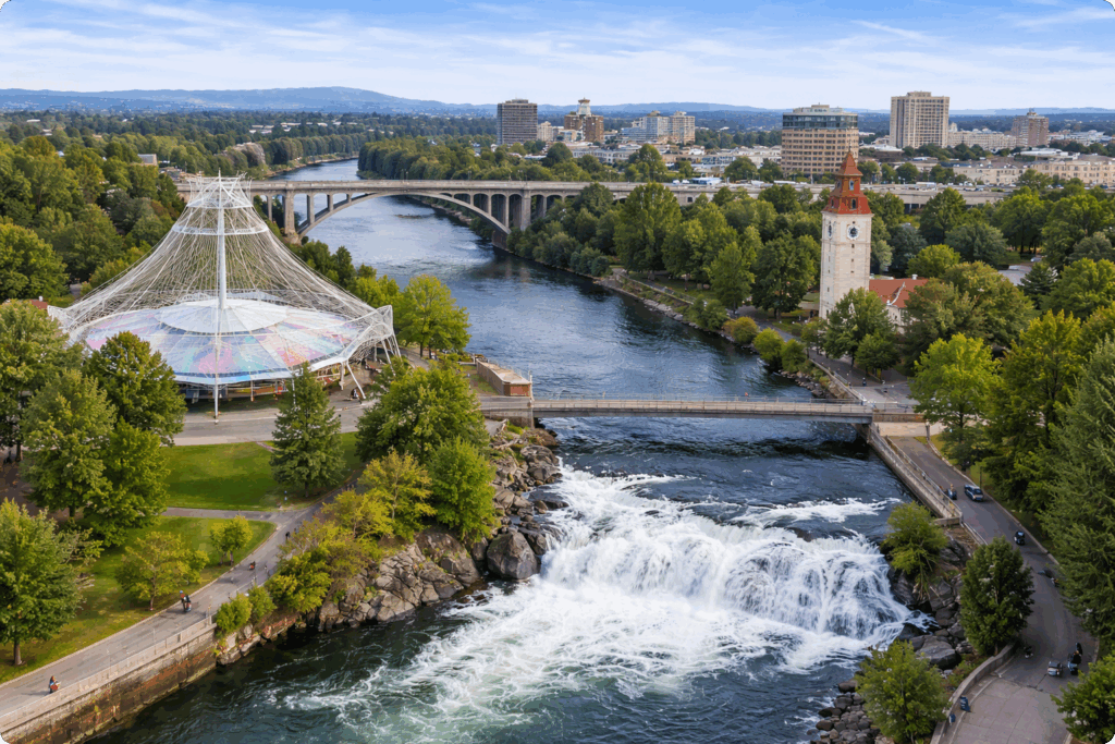 Aerial view of Spokane Falls on the Spokane River in downtown Spokane, Washington, with Riverfront Park pavilion, historic clock tower, and surrounding city skyline.