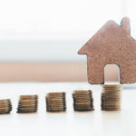Stack of coins with a house made of cardboard in the background.