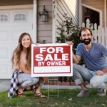Man and woman posing with a for sale sign in front of their house