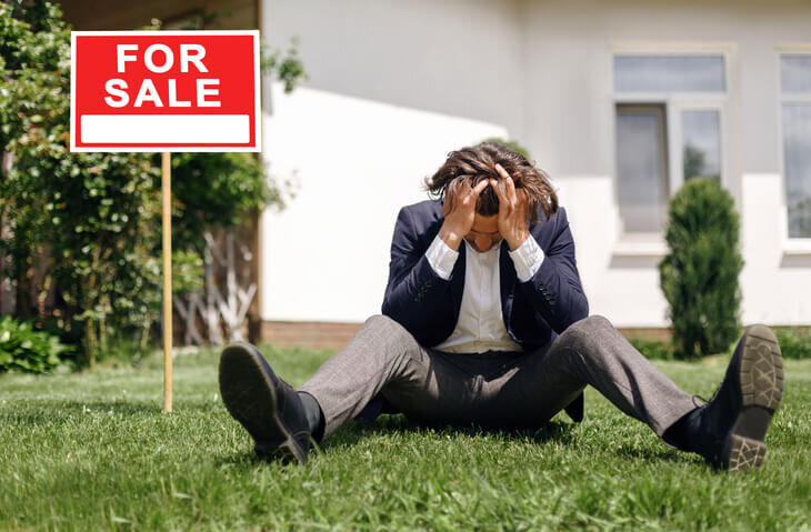 Depressed-real-estate-agent-sitting-on-grass-in-front-of-house-for-sale-failed-home-sale-bridgetown-home-buyers