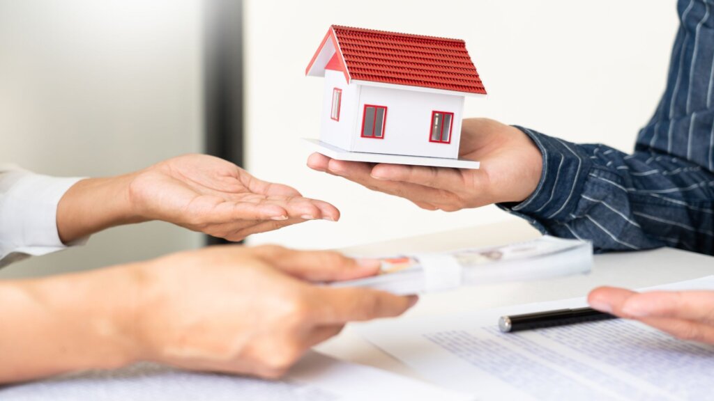 Two pair of hands, with one of them holding a miniature house, giving it to the other person