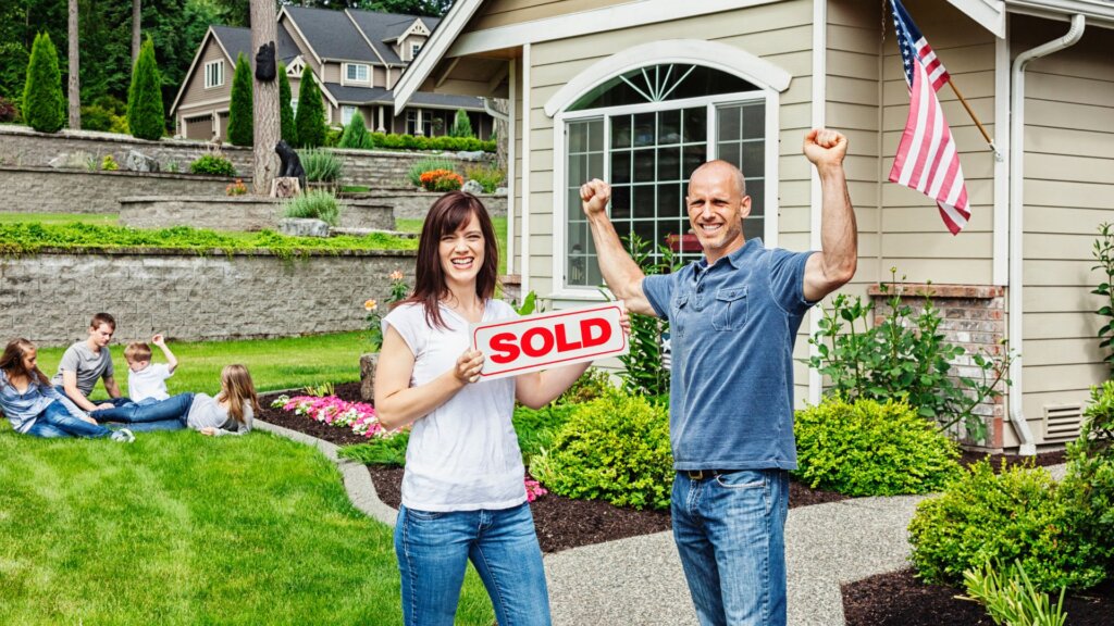 A couple in front of a house with the lady holding a signboard that says "Sold"