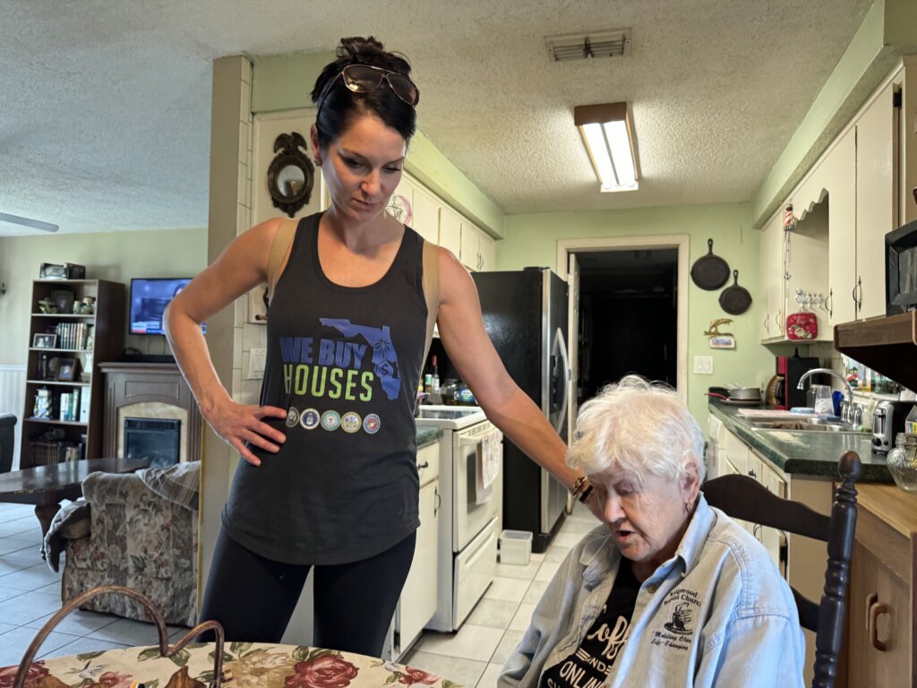 Kady Andreoli, a real estate professional with ‘We Buy Houses’ in Florida, stands in a kitchen, offering support by placing a hand on the shoulder of an elderly woman seated at the table. Wearing a branded tank top, Kady conveys care and professionalism. The setting is a cozy, lived-in home with a fireplace, bookshelves, and a vintage-style kitchen, reflecting a personal and compassionate approach to helping homeowners in need.