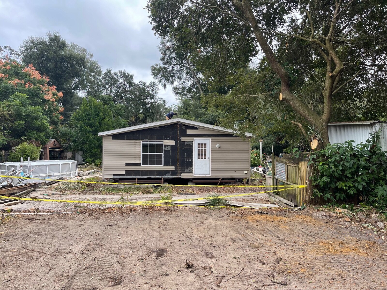 A mobile home mid-rehab on private land in Florida, siding partially stripped, with a worker on the roof