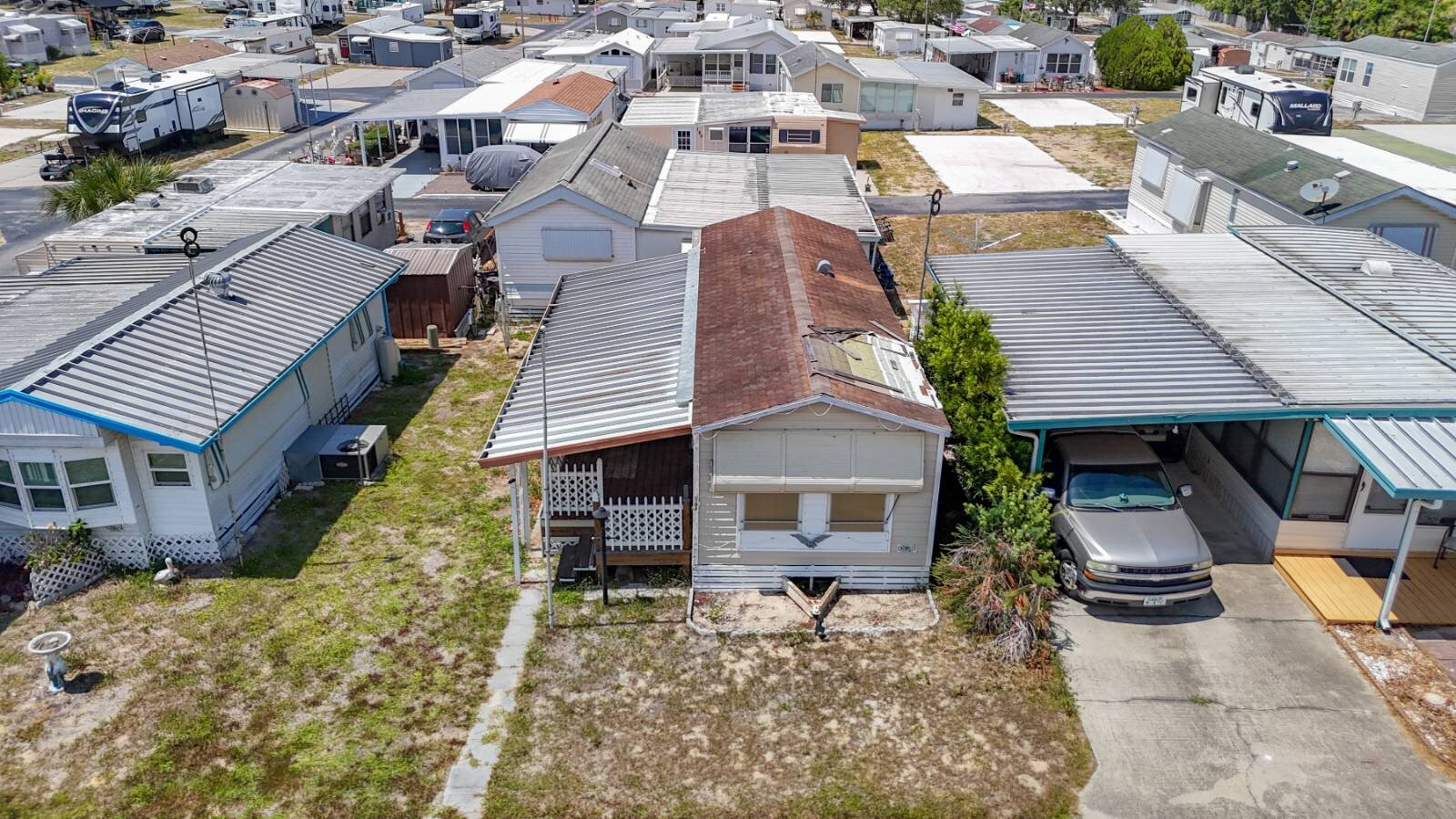 Aerial view of an older Florida mobile home on a rented lot in a Hudson mobile home park, its roof patched and failing, with neighbouring homes close on both sides