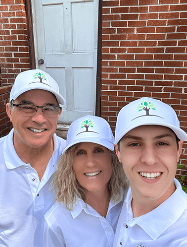 Stephen, Cynthia and Anthony standing in front of a fixer upper brick house with company hats and shirts on.