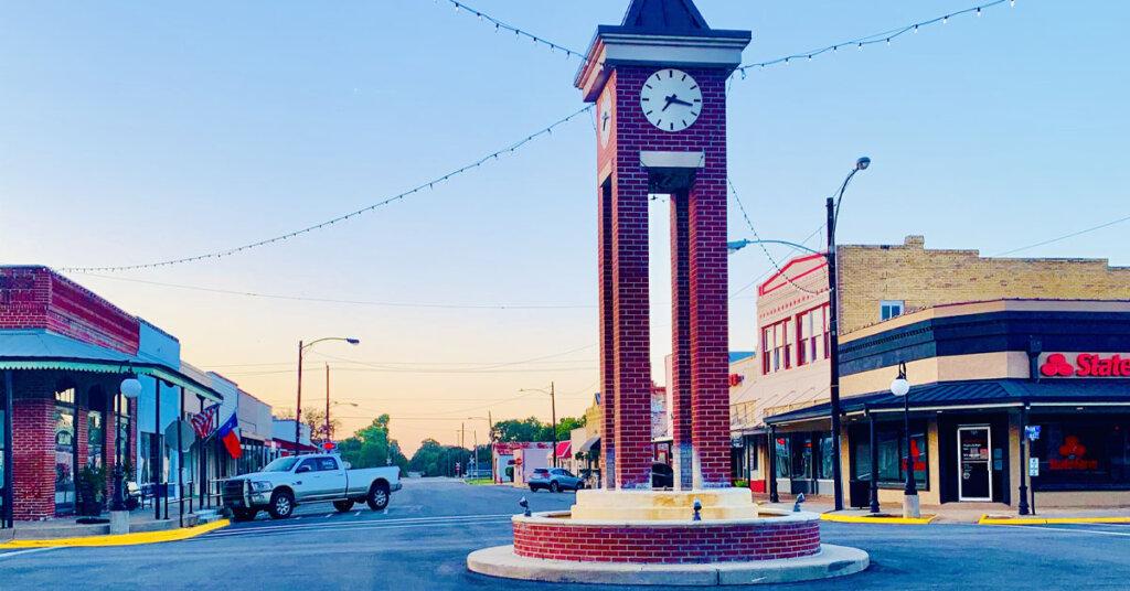 picture of the clock in downtown Sealy, Texas. The clock is in the center of an intersection and there are retail stores lining down the street from the clock.