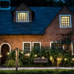 A rustic two-story brick house at dusk with a dark blue shingle roof, glowing windows, an arched wooden front door, and a landscaped garden illuminated by warm string lights.