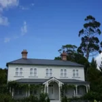 large, two-story white heritage house with a dark hipped roof, brick chimneys, decorative arched windows, and a covered front veranda surrounded by lush greenery beneath a blue sky.