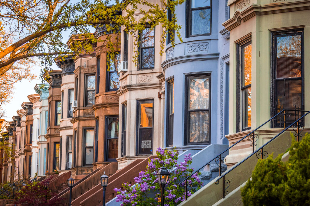 A row of brownstone houses, each house is a different color with a long set of steps leading up to the main enterance of the 3 story building. Pretty foliage and street lamp surrounding.