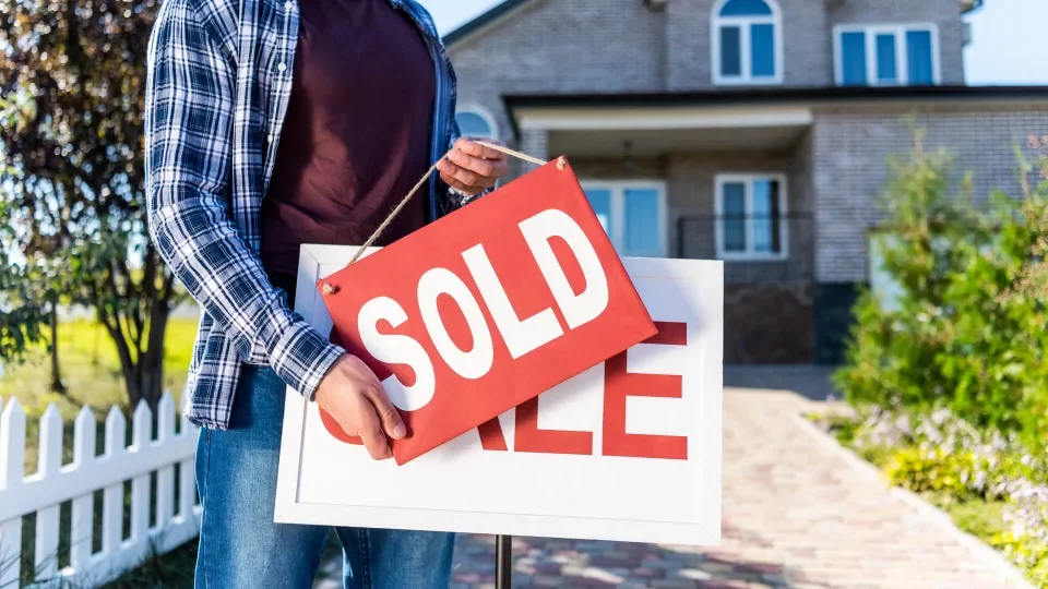 Close up photo of a man standing in front of a house holding a red and white SOLD sign.