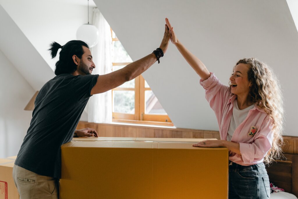 A white man with a beard and mustache, black t-shirt and beige cargo pants and a white women with blond curly hair, pink sweater and blue jeans are smiling and giving each other a high five, while standing over a moving box in front of a window.