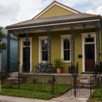 A modest New Orleans double-shotgun rental home with a For Sale sign in the yard, tenant belongings visible on the porch