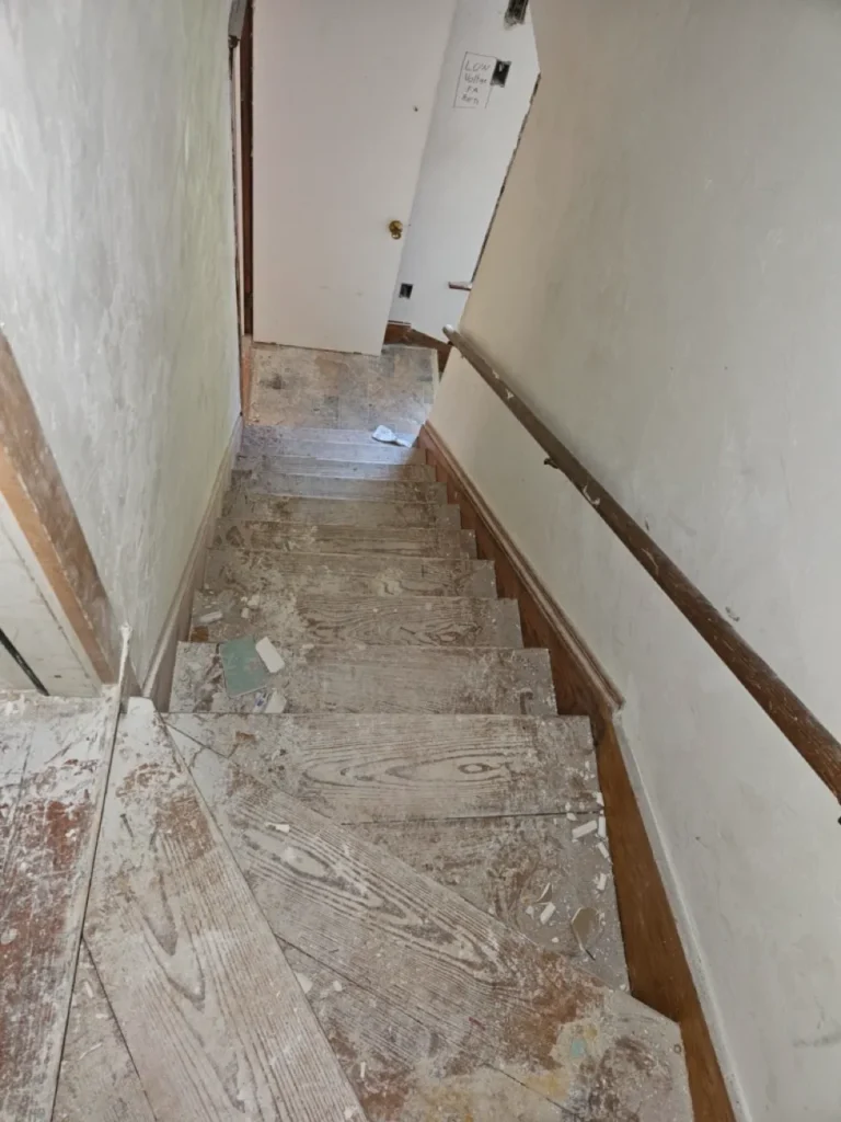 Deteriorated wooden interior staircase covered in drywall dust and construction debris inside a distressed house.