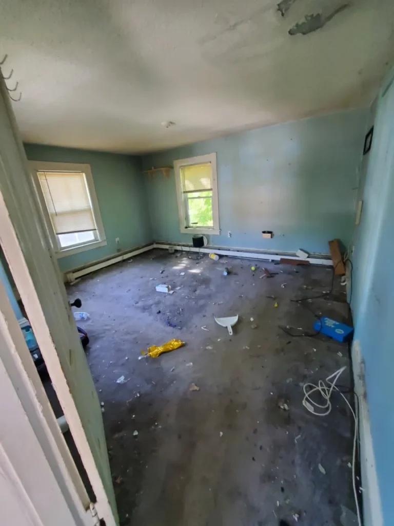 Neglected upstairs bedroom with littered flooring, stained walls, and peeling ceiling paint in a vacant house.