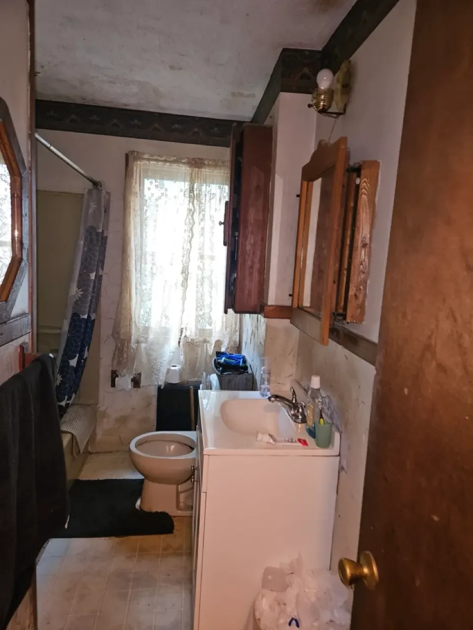 Outdated bathroom in a Greater Boston property featuring worn walls, a vintage vanity, and lace curtains