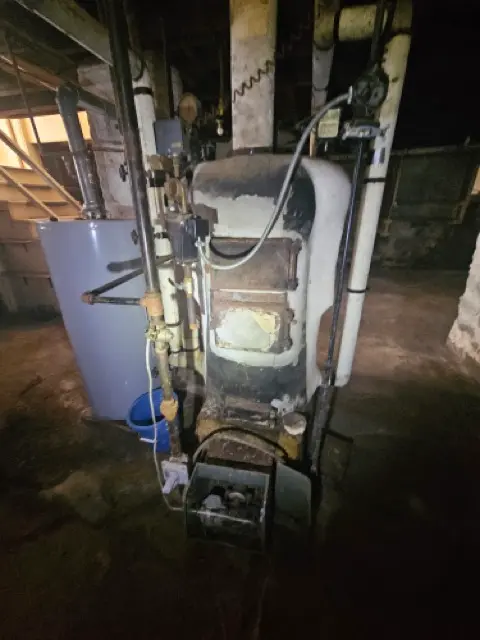 Dark basement with an old, weathered oil or gas boiler with peeling insulation, wrapped pipes, and an adjacent water heater that needs servicing or replacement.