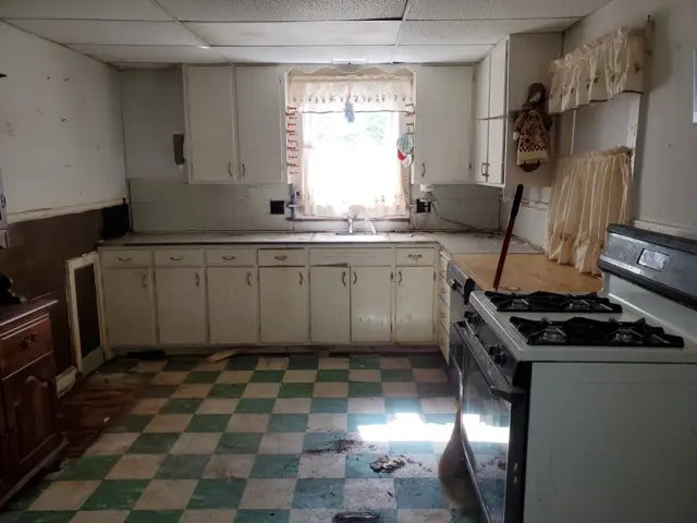 Outdated kitchen interior with damaged green and white checkered tile floor, old white cabinets, and a window above the sink.