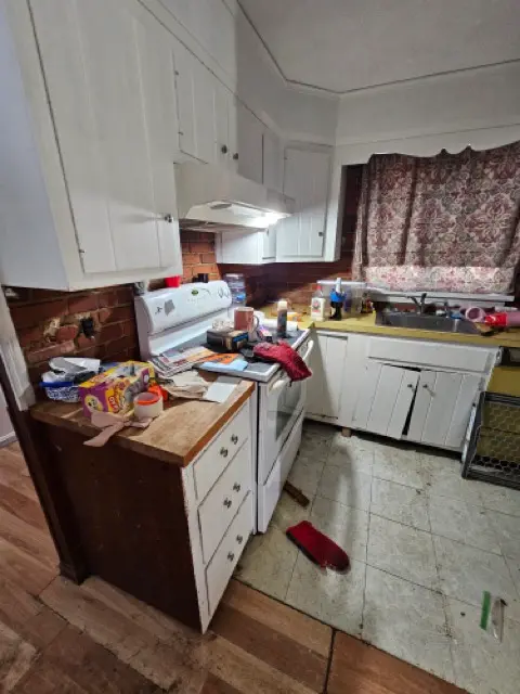 Cluttered kitchen with white cabinets, featuring a broken lower cabinet door hanging off its hinges, and a section of missing drywall and a heavily stained floor.