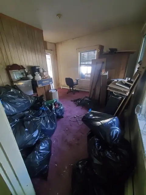 Bedroom space with purple carpeting filled with large black trash bags and stacked wood panels.
