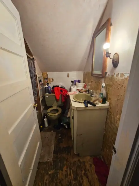 Small, cluttered bathroom with a slanted ceiling and green fixtures, showing heavily peeling wall coverings and damaged plaster that require extensive repair.