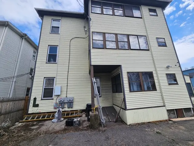 Multi-story apartment building with light-yellow siding, multiple dark-framed windows, and utility meters on the lower exterior wall, showing signs of wear that require maintenance and repair.