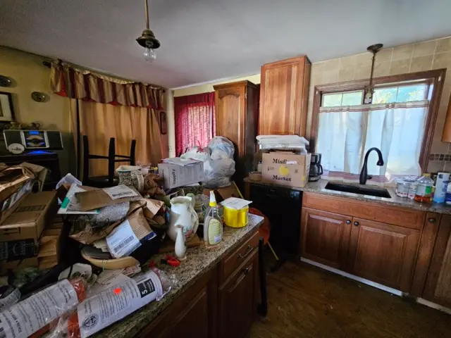 Kitchen counter and island piled high with cardboard boxes, mail, and household supplies.