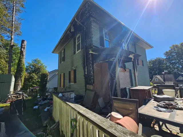 Exterior view of a two-story house with green siding, showing severe fire and smoke damage.