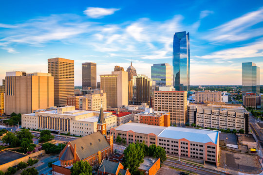 A sunny daytime skyline of Oklahoma City with modern high-rise buildings, historic architecture, and a bright blue sky with clouds.