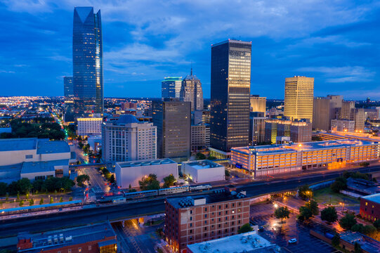 A vibrant cityscape of Oklahoma City at twilight, featuring brightly lit skyscrapers and downtown buildings under a deep blue sky.