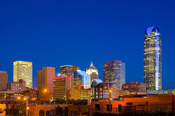 A nighttime view of Oklahoma City with its illuminated skyline, including iconic buildings and a clear dark blue backdrop.