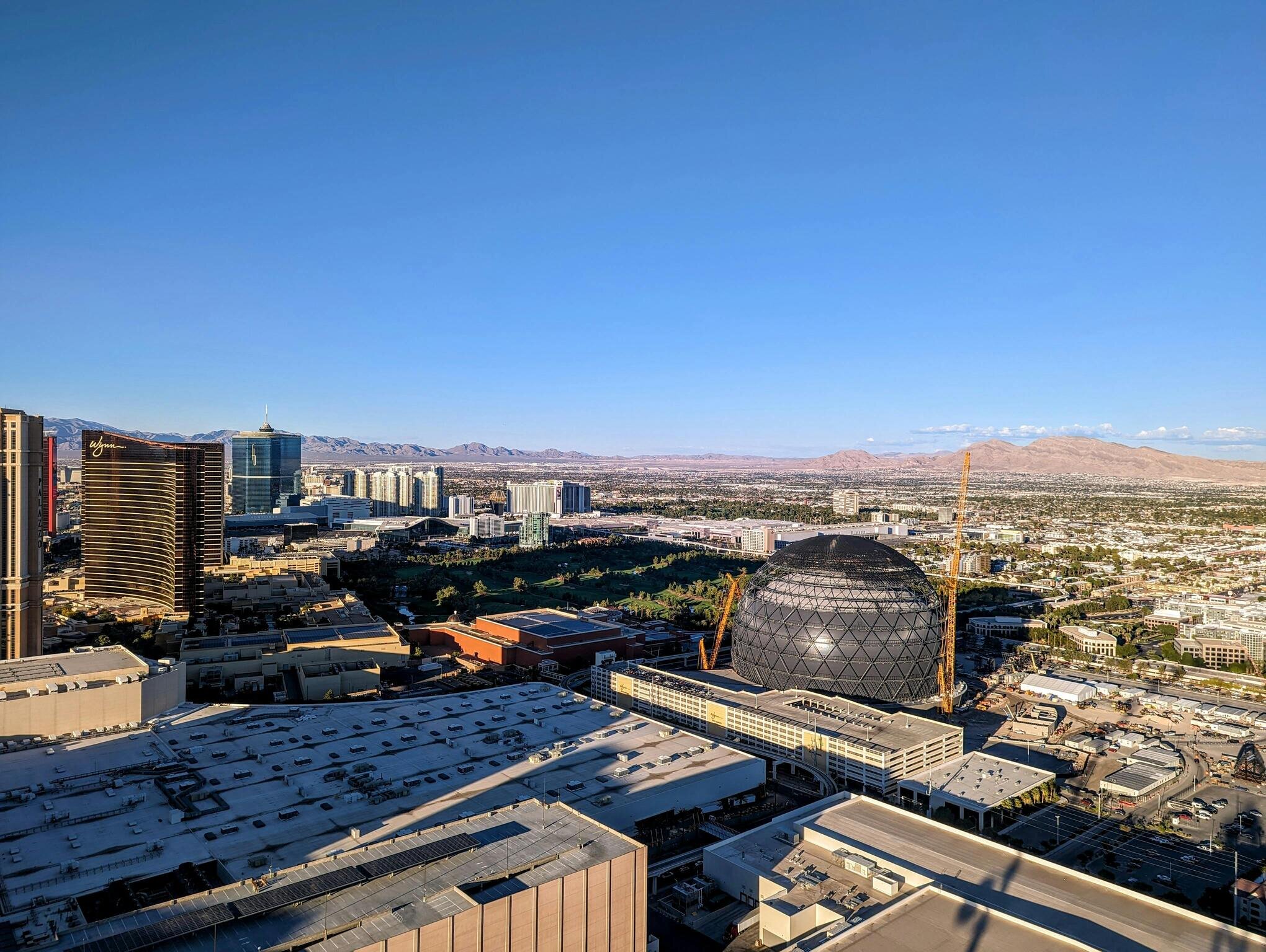 A cityscape with modern buildings, a large spherical structure, and mountains in the background under a clear blue sky.