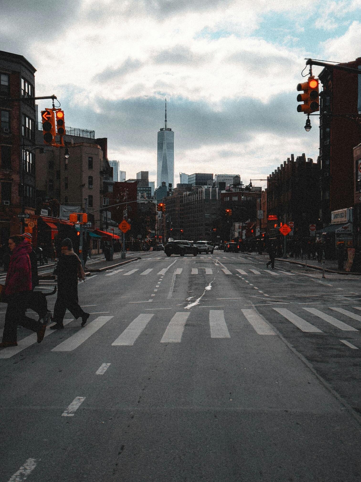 A city street with pedestrians crossing, traffic lights, and the One World Trade Center visible in the background under a cloudy sky.