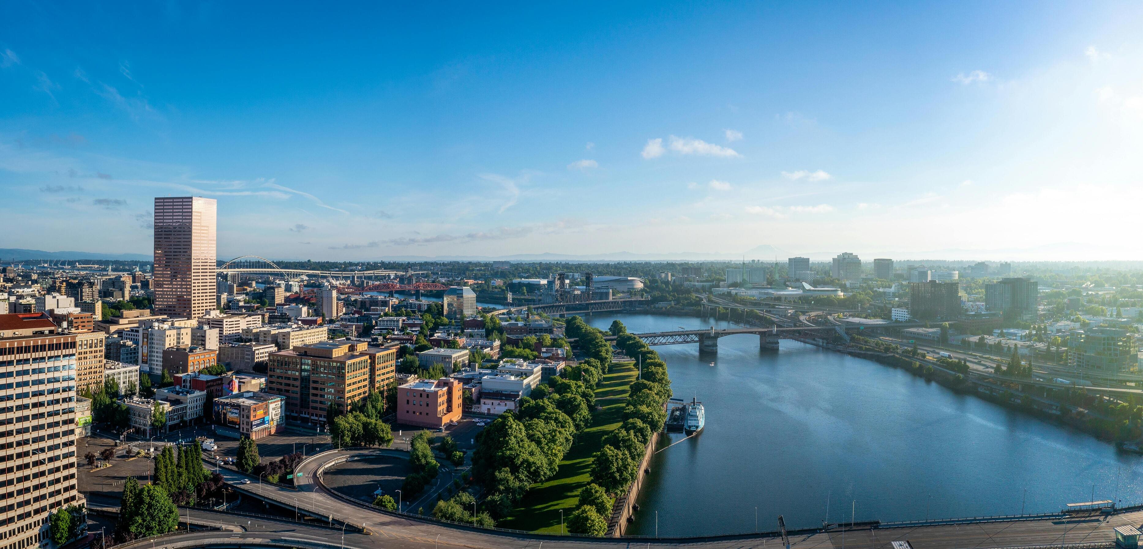 A panoramic view of a city with a prominent skyscraper, bridges crossing a calm river, and tree-lined parks under a bright blue sky.