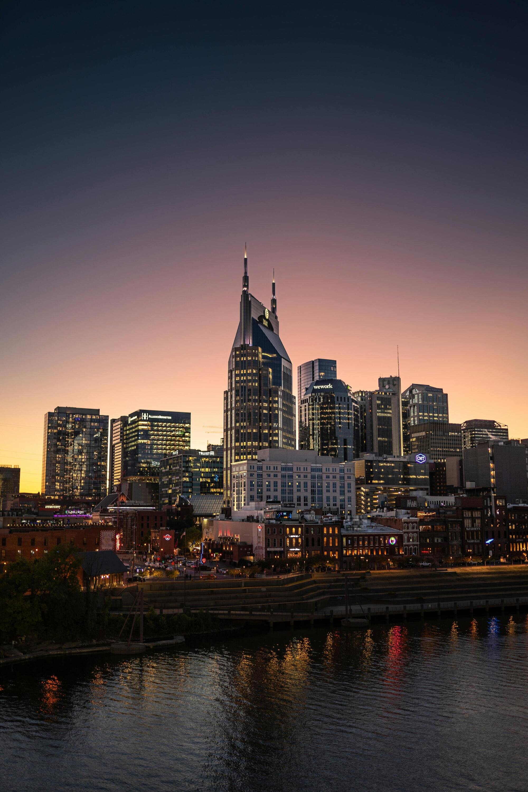 Nashville skyline at dusk, featuring the iconic AT&T building and surrounding modern high-rises with reflections on the river.
